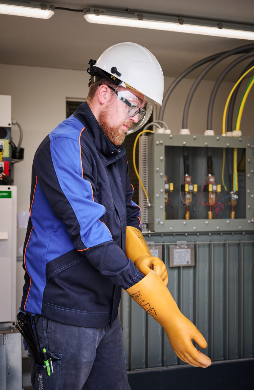Ein Elektriker in Schutzkleidung und Helm zieht gelbe Isolierhandschuhe an vor einem Schaltschrank mit Elektrokabeln in der Sporthalle Schönefeld Eupen. Die Szene zeigt die elektrotechnische Ausstattung der barrierefreien Mehrzweckhalle, die mit moderner Sicherheitstechnik, intelligenter LED-Beleuchtung, IT-Verkabelung und einer Photovoltaikanlage ausgestattet ist.
