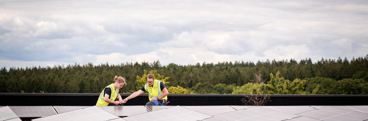 Auf dem Dach der Sporthalle Schönefeld Eupen arbeiten zwei Techniker an der Photovoltaikanlage, die Teil der modernen elektrotechnischen Ausstattung der barrierefreien Mehrzweckhalle ist. Die Installation unterstützt eine intelligente LED-Beleuchtung, IT-Verkabelung und umfassende Sicherheitstechnik.
