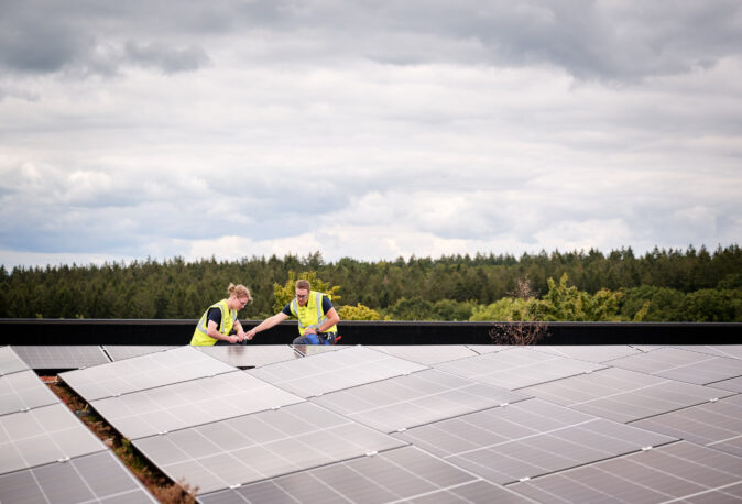 Auf dem Dach der Sporthalle Schönefeld Eupen arbeiten zwei Techniker an der Photovoltaikanlage, die Teil der modernen elektrotechnischen Ausstattung der barrierefreien Mehrzweckhalle ist. Die Installation unterstützt eine intelligente LED-Beleuchtung, IT-Verkabelung und umfassende Sicherheitstechnik.