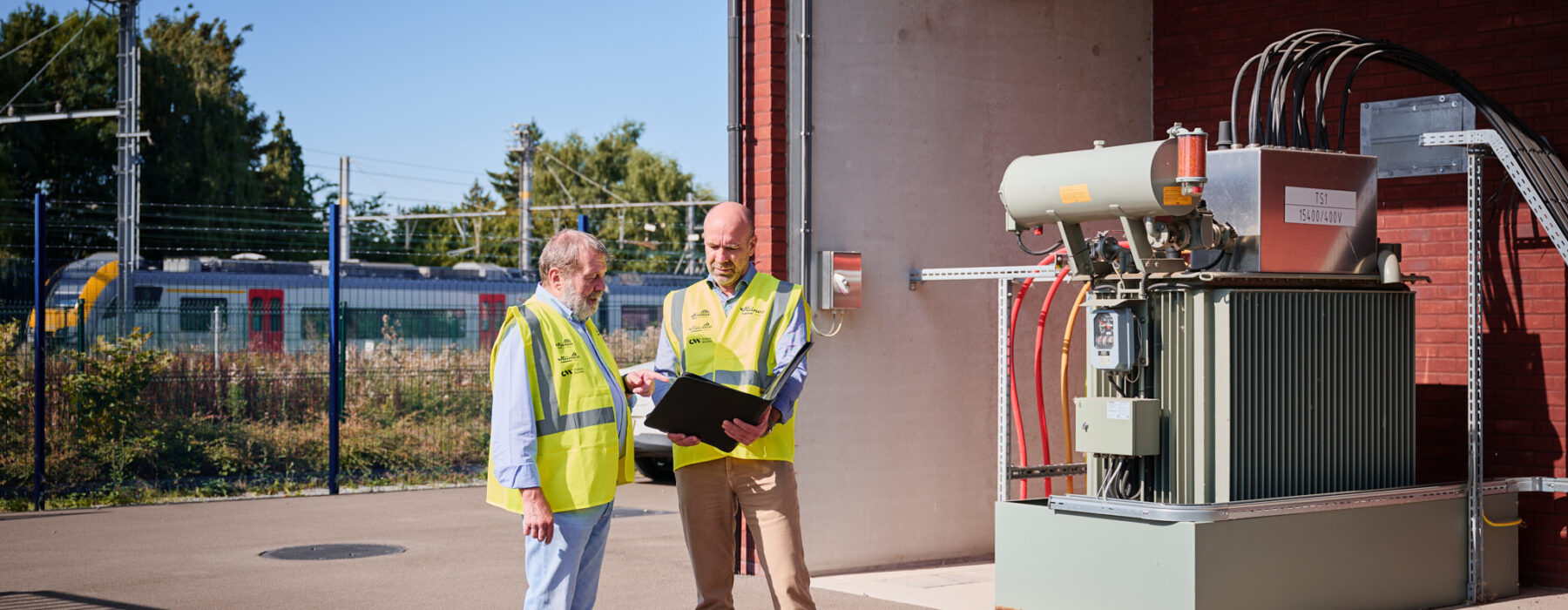 Zwei Techniker in Warnwesten besprechen vor einer Mittelspannungsstation mit Transformator-Gleichrichter-Gruppen die Planung und den Bau eines Infrabel Gleichrichterunterwerks in Lüttich zur Stromversorgung Schienenverkehr und Gleichstrom Bahnenergie. Im Hintergrund ist ein Zug und eine Metallstation sichtbar.