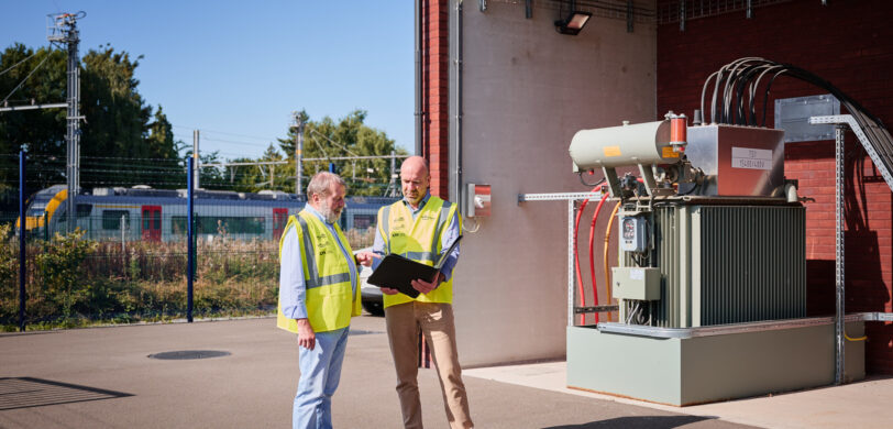 Zwei Techniker in Warnwesten besprechen vor einer Mittelspannungsstation mit Transformator-Gleichrichter-Gruppen die Planung und den Bau eines Infrabel Gleichrichterunterwerks in Lüttich zur Stromversorgung Schienenverkehr und Gleichstrom Bahnenergie. Im Hintergrund ist ein Zug und eine Metallstation sichtbar.