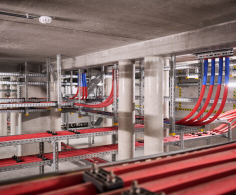 Innenansicht eines Infrabel Gleichrichterunterwerks in Lüttich mit zahlreichen roten und blauen Kabeln, die auf Metalltrassen verlegt sind. Die Mittelspannungsstation zeigt die sorgfältige Planung und den Bau von Bahnunterwerken, wobei Transformator-Gleichrichter-Gruppen in einer robusten Metallstation installiert sind. Diese Anlage dient der Stromversorgung im Schienenverkehr und gewährleistet die zuverlässige Bereitstellung von Gleichstrom Bahnenergie.