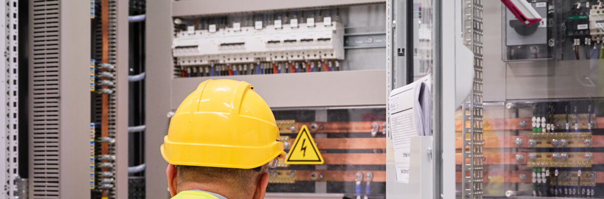 Ein Techniker mit gelbem Helm und Warnweste inspiziert ein Infrabel Gleichrichterunterwerk in Lüttich. Im Hintergrund sind Mittelspannungsstationen mit Transformator-Gleichrichter-Gruppen und Schaltanlagen zu sehen, die für die Stromversorgung im Schienenverkehr und die Gleichstrom Bahnenergie verantwortlich sind. Die Metallstation zeigt die komplexe Planung und den Bau von Bahnunterwerken.