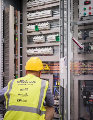 Ein Techniker mit gelbem Helm und Warnweste inspiziert ein Infrabel Gleichrichterunterwerk in Lüttich. Im Hintergrund sind Mittelspannungsstationen mit Transformator-Gleichrichter-Gruppen und Schaltanlagen zu sehen, die für die Stromversorgung im Schienenverkehr und die Gleichstrom Bahnenergie verantwortlich sind. Die Metallstation zeigt die komplexe Planung und den Bau von Bahnunterwerken.