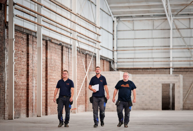 Drei Elektriker im Industrie Bereich gehen durch eine große Halle mit unverputzten Backstein- und Betonwänden, bereit für Elektro Arbeiten an Schalttafeln, Schaltschränken, Mittel- und Hochspannungsanlagen sowie Verkabelungssystemen und Glasfaserarbeiten im Kontext von Erneuerbaren Energien.