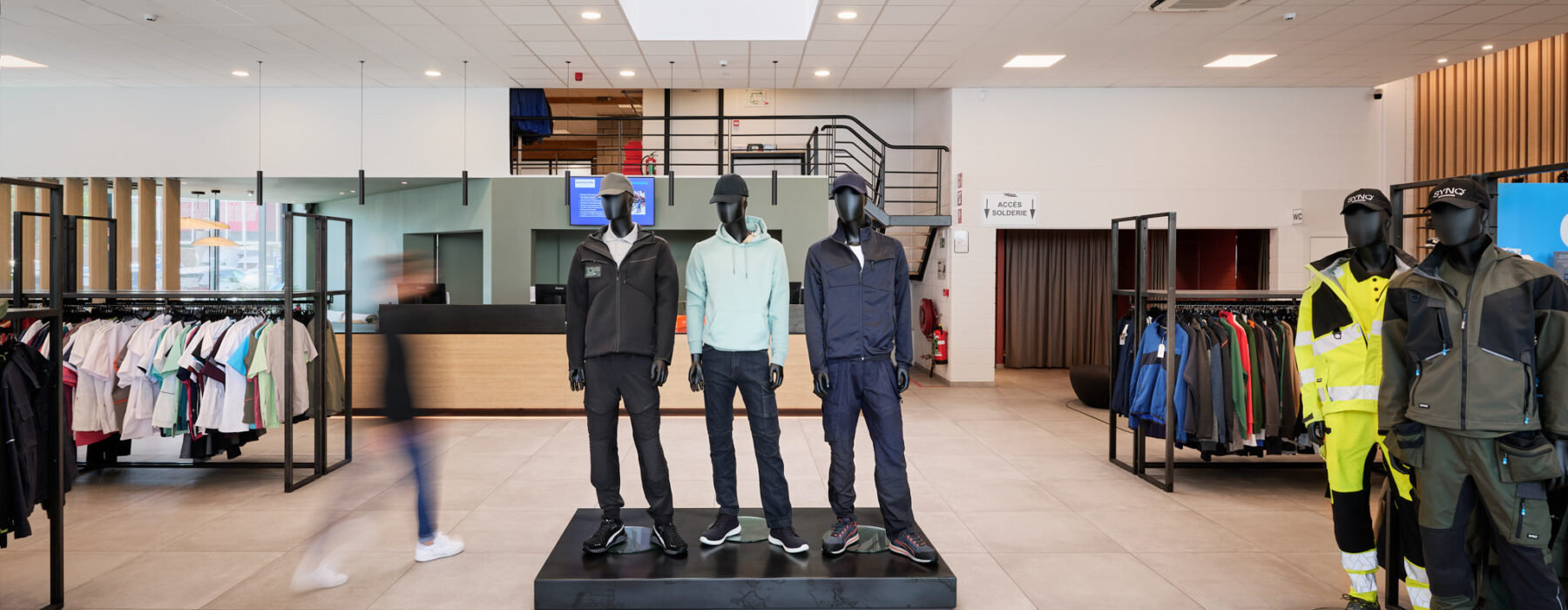 Ein moderner, gut beleuchteter Verkaufsraum mit mehreren Schaufensterpuppen, die Arbeits- und Schutzkleidung präsentieren. Im Hintergrund sind Kleiderständer mit verschiedenen Berufskleidungsstücken zu sehen. Die klare Raumgestaltung und Beleuchtung deuten auf eine professionelle industrielle Elektroinstallation für die Industrie hin, die auch Aspekte wie Neubau und Inbetriebnahme Schaltschränke sowie elektrotechnische Sanierung und Wiederherstellung der Stromversorgung berücksichtigt. Der Raum könnte Teil eines Wiederaufbaus Produktionsstandort nach Umweltkatastrophe sein, bei dem unter Zeitdruck eine elektrotechnische Wiederherstellung nach Zerstörung und die Rekonstruktion von Mittelspannung und Niederspannungsverteilung erfolgt.