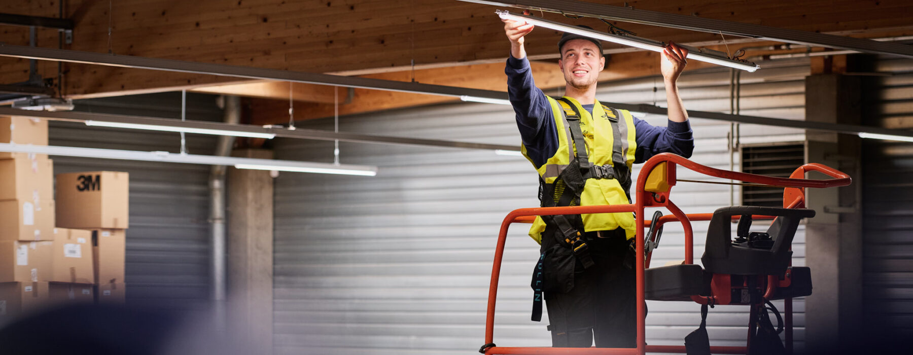 Ein Elektriker führt eine industrielle Elektroinstallation für die Industrie durch und erneuert Beleuchtungssysteme in einer Halle. Er steht auf einer Hebebühne und arbeitet unter Zeitdruck an der Wiederherstellung der Stromversorgung. Die Szene symbolisiert den Wiederaufbau und die elektrotechnische Sanierung eines Produktionsstandorts nach Zerstörung, inklusive Neubau und Inbetriebnahme von Schaltschränken sowie Rekonstruktion von Mittelspannung und Niederspannungsverteilung. Die Notversorgung und elektrotechnische Wiederherstellung nach Umweltkatastrophe sind zentrale Aspekte dieses Einsatzes.