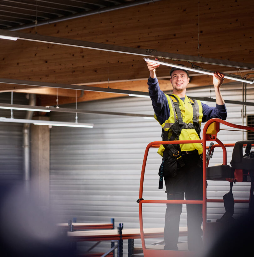 Ein Elektriker führt eine industrielle Elektroinstallation für die Industrie durch und erneuert Beleuchtungssysteme in einer Halle. Er steht auf einer Hebebühne und arbeitet unter Zeitdruck an der Wiederherstellung der Stromversorgung. Die Szene symbolisiert den Wiederaufbau und die elektrotechnische Sanierung eines Produktionsstandorts nach Zerstörung, inklusive Neubau und Inbetriebnahme von Schaltschränken sowie Rekonstruktion von Mittelspannung und Niederspannungsverteilung. Die Notversorgung und elektrotechnische Wiederherstellung nach Umweltkatastrophe sind zentrale Aspekte dieses Einsatzes.