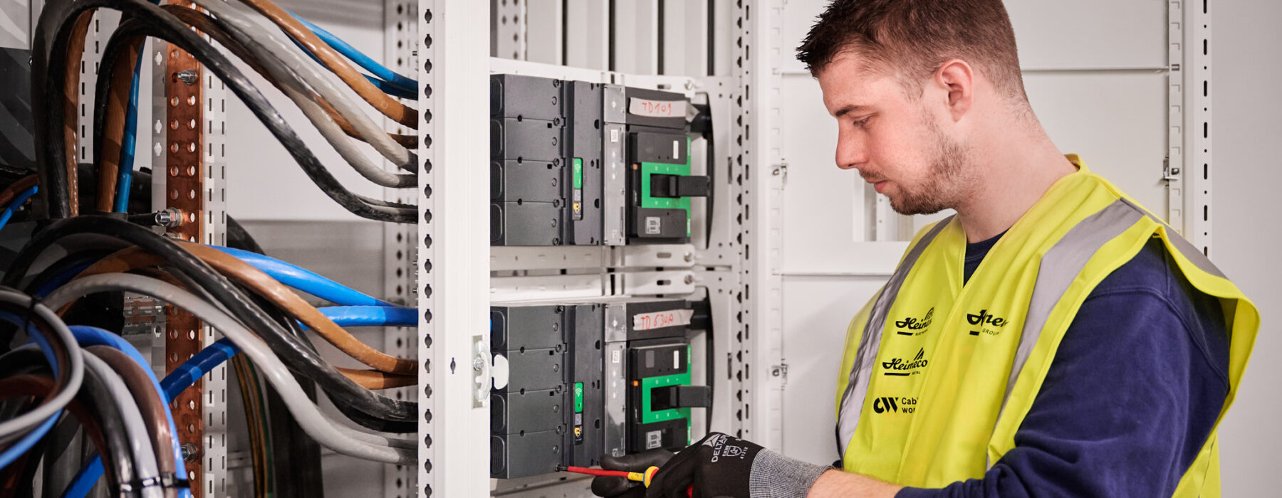 Ein Elektriker führt die industrielle Elektroinstallation unter Zeitdruck durch und arbeitet am Neubau und der Inbetriebnahme von Schaltschränken. Dabei erfolgt die elektrotechnische Sanierung und Wiederherstellung der Stromversorgung in einem Produktionsstandort. Die Tätigkeit umfasst die Rekonstruktion von Mittelspannung und Niederspannungsverteilung sowie die Erneuerung von Mittelspannungsanlagen. Dies ist Teil der elektrotechnischen Wiederherstellung nach Zerstörung und des Wiederaufbaus nach einer Umweltkatastrophe, inklusive Notversorgung.