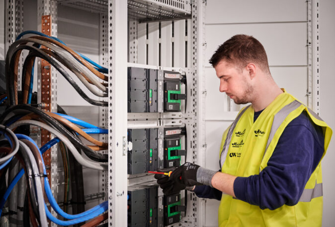 Ein Elektriker führt die industrielle Elektroinstallation unter Zeitdruck durch und arbeitet am Neubau und der Inbetriebnahme von Schaltschränken. Dabei erfolgt die elektrotechnische Sanierung und Wiederherstellung der Stromversorgung in einem Produktionsstandort. Die Tätigkeit umfasst die Rekonstruktion von Mittelspannung und Niederspannungsverteilung sowie die Erneuerung von Mittelspannungsanlagen. Dies ist Teil der elektrotechnischen Wiederherstellung nach Zerstörung und des Wiederaufbaus nach einer Umweltkatastrophe, inklusive Notversorgung.