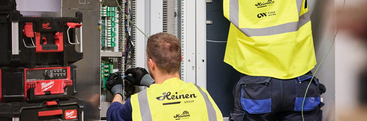 Zwei Elektriker in gelben Warnwesten führen eine industrielle Elektroinstallation durch. Sie erneuern und rekonstruieren Mittelspannungsanlagen in einem Schaltschrank, um die Wiederherstellung der Stromversorgung und Notversorgung sicherzustellen. Die Arbeiten erfolgen unter Zeitdruck im Rahmen der elektrotechnischen Sanierung und Wiederherstellung nach Zerstörung, beispielsweise beim Wiederaufbau eines Produktionsstandorts nach einer Umweltkatastrophe. Werkzeugkoffer sind neben dem Schaltschrank sichtbar, während die Techniker Kabel verlegen und Schaltschränke neu bauen und in Betrieb nehmen.