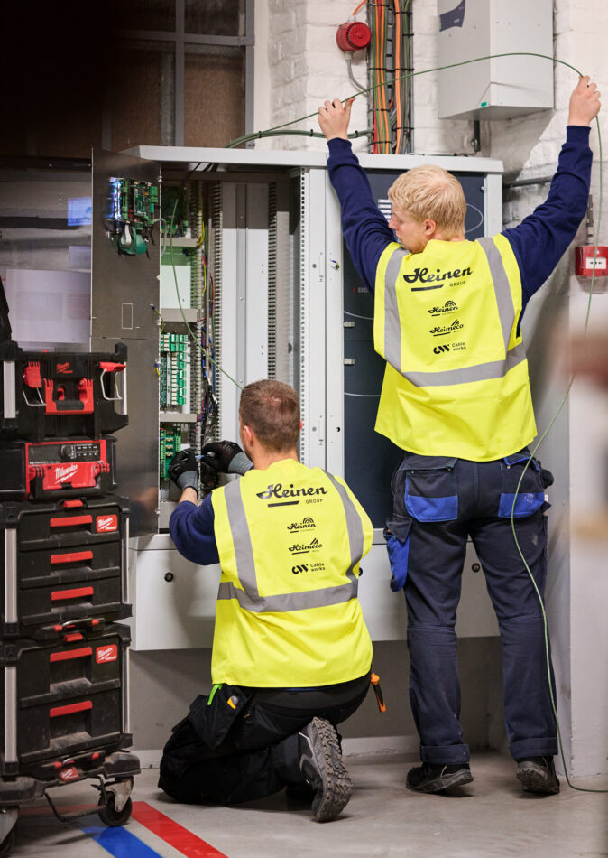 Zwei Elektriker in gelben Warnwesten führen eine industrielle Elektroinstallation durch. Sie erneuern und rekonstruieren Mittelspannungsanlagen in einem Schaltschrank, um die Wiederherstellung der Stromversorgung und Notversorgung sicherzustellen. Die Arbeiten erfolgen unter Zeitdruck im Rahmen der elektrotechnischen Sanierung und Wiederherstellung nach Zerstörung, beispielsweise beim Wiederaufbau eines Produktionsstandorts nach einer Umweltkatastrophe. Werkzeugkoffer sind neben dem Schaltschrank sichtbar, während die Techniker Kabel verlegen und Schaltschränke neu bauen und in Betrieb nehmen.