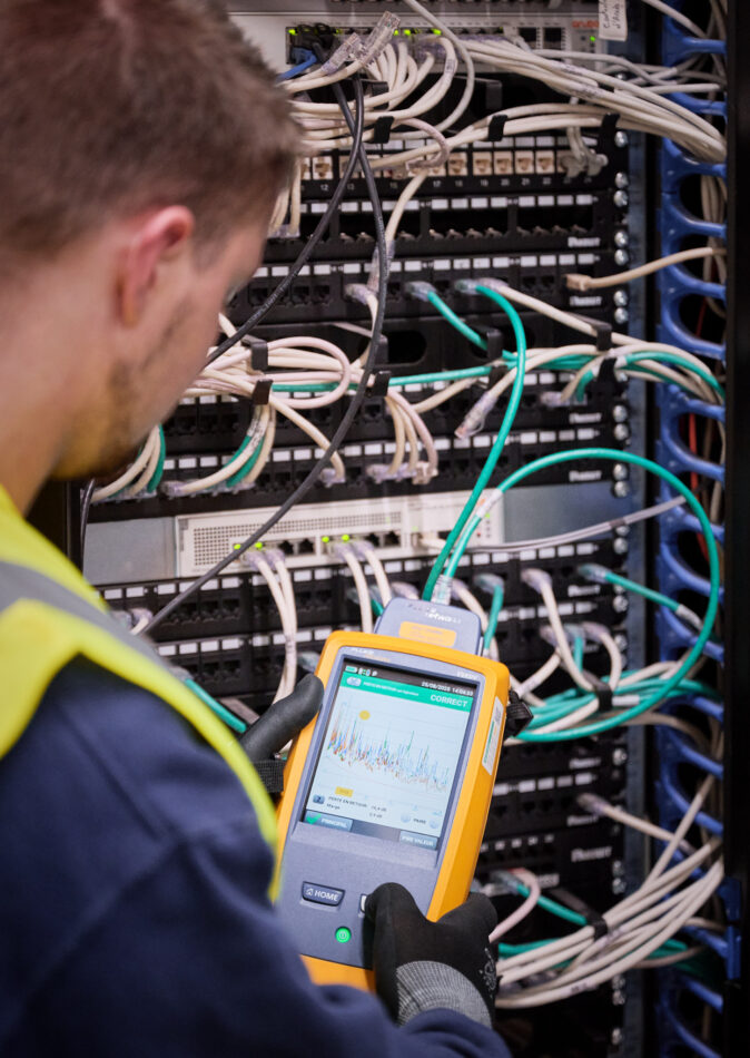 Ein Techniker führt eine Messung an einer komplexen Verkabelung in einem Schaltschrank durch. Die Installation zeigt zahlreiche weiße und grüne Kabel, die in Patchpanels und Netzwerkgeräte eingesteckt sind. Der Techniker trägt Handschuhe und eine Sicherheitsweste, was auf eine industrielle Umgebung hinweist. Diese Szene steht exemplarisch für die Elektroinstallation für die Industrie, insbesondere im Rahmen der Erneuerung von Mittelspannungsanlagen und der elektrotechnischen Wiederherstellung nach Zerstörung. Der Fokus liegt auf der Wiederherstellung der Stromversorgung und der Notversorgung, die bei der Rekonstruktion von Mittelspannung und Niederspannungsverteilung sowie der elektrotechnischen Sanierung und dem Wiederaufbau eines Produktionsstandorts nach einer Umweltkatastrophe unter Zeitdruck erfolgen muss. Der Neubau und die Inbetriebnahme von Schaltschränken sind ebenfalls Teil dieser anspruchsvollen industriellen Elektroinstallation.