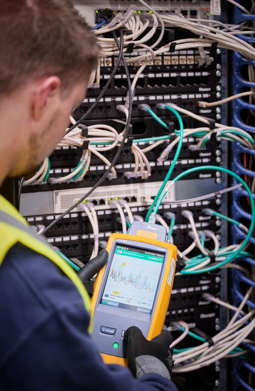 Ein Techniker führt eine Messung an einer komplexen Verkabelung in einem Schaltschrank durch. Die Installation zeigt zahlreiche weiße und grüne Kabel, die in Patchpanels und Netzwerkgeräte eingesteckt sind. Der Techniker trägt Handschuhe und eine Sicherheitsweste, was auf eine industrielle Umgebung hinweist. Diese Szene steht exemplarisch für die Elektroinstallation für die Industrie, insbesondere im Rahmen der Erneuerung von Mittelspannungsanlagen und der elektrotechnischen Wiederherstellung nach Zerstörung. Der Fokus liegt auf der Wiederherstellung der Stromversorgung und der Notversorgung, die bei der Rekonstruktion von Mittelspannung und Niederspannungsverteilung sowie der elektrotechnischen Sanierung und dem Wiederaufbau eines Produktionsstandorts nach einer Umweltkatastrophe unter Zeitdruck erfolgen muss. Der Neubau und die Inbetriebnahme von Schaltschränken sind ebenfalls Teil dieser anspruchsvollen industriellen Elektroinstallation.