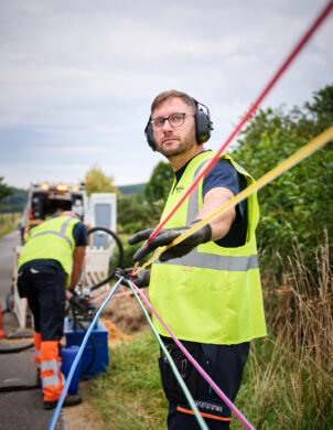 Zwei Techniker mit Schutzkleidung und Gehörschutz arbeiten an der Stromnetzinfrastruktur in Belgien. Sie ziehen farbige Kabel für Mittelspannungsanlagen und Niederspannungsverteilung, die Teil des Neubaus und Umbaus von schlüsselfertigen Trafostationen für die Verteilnetzbetreiber ORES und RESA sind. Im Hintergrund ist ein Servicefahrzeug zu sehen, das für die Energieinfrastruktur Belgien eingesetzt wird. Die Arbeiten umfassen auch Transformatoren und Beton-Fertigstationen.