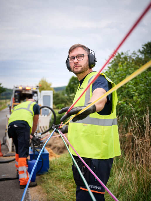 Zwei Techniker mit Schutzkleidung und Gehörschutz arbeiten an der Stromnetzinfrastruktur in Belgien. Sie ziehen farbige Kabel für Mittelspannungsanlagen und Niederspannungsverteilung, die Teil des Neubaus und Umbaus von schlüsselfertigen Trafostationen für die Verteilnetzbetreiber ORES und RESA sind. Im Hintergrund ist ein Servicefahrzeug zu sehen, das für die Energieinfrastruktur Belgien eingesetzt wird. Die Arbeiten umfassen auch Transformatoren und Beton-Fertigstationen.
