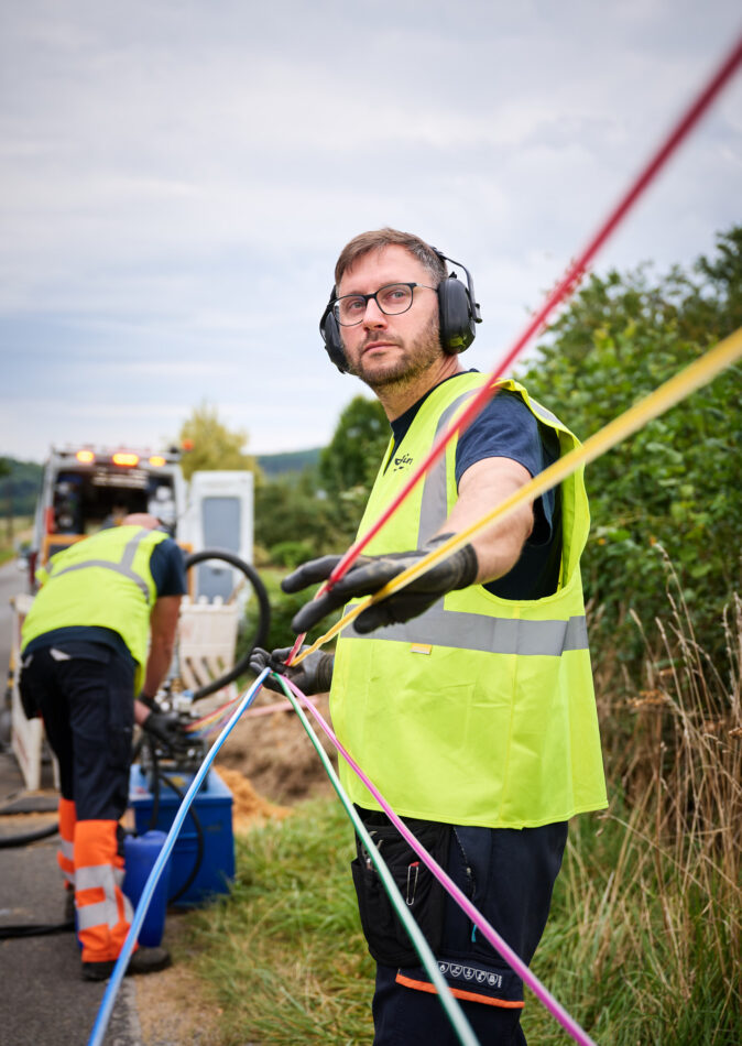 Zwei Techniker mit Schutzkleidung und Gehörschutz arbeiten an der Stromnetzinfrastruktur in Belgien. Sie ziehen farbige Kabel für Mittelspannungsanlagen und Niederspannungsverteilung, die Teil des Neubaus und Umbaus von schlüsselfertigen Trafostationen für die Verteilnetzbetreiber ORES und RESA sind. Im Hintergrund ist ein Servicefahrzeug zu sehen, das für die Energieinfrastruktur Belgien eingesetzt wird. Die Arbeiten umfassen auch Transformatoren und Beton-Fertigstationen.