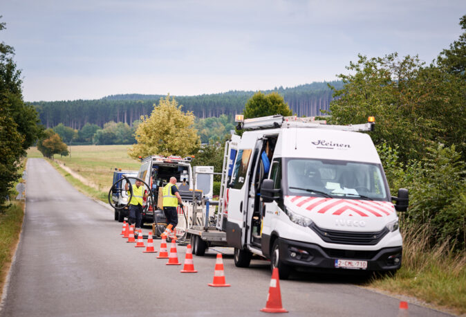 Zwei Techniker in Warnwesten arbeiten an der Stromnetzinfrastruktur am Straßenrand in ländlicher Umgebung. Vor ihnen stehen zwei Transporter mit geöffneter Ladeklappe, aus denen Kabel und Werkzeuge entnommen werden. Die Szene zeigt den Neubau und Umbau von Mittelspannungsanlagen und Niederspannungsverteilung für Verteilnetzbetreiber ORES und RESA in Belgien. Die Arbeiten umfassen schlüsselfertige Trafostationen und den Einsatz von Transformatoren sowie Beton-Fertigstationen zur Modernisierung der Energieinfrastruktur Belgien.