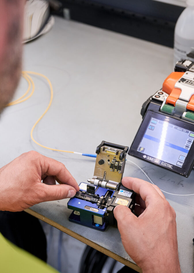 Ein Techniker arbeitet an einer Fusionsspleißmaschine zur Verbindung von Glasfaserkabeln, ein wichtiger Schritt für die Stromnetzinfrastruktur und Mittelspannungsanlagen bei Verteilnetzbetreibern ORES und RESA. Die präzise Handhabung unterstützt den Neubau und Umbau von Trafostationen sowie die Installation schlüsselfertiger Trafostationen und Beton-Fertigstationen in der Energieinfrastruktur Belgien. Werkzeuge und Geräte zur Niederspannungsverteilung und für Transformatoren sind auf dem Arbeitstisch sichtbar.