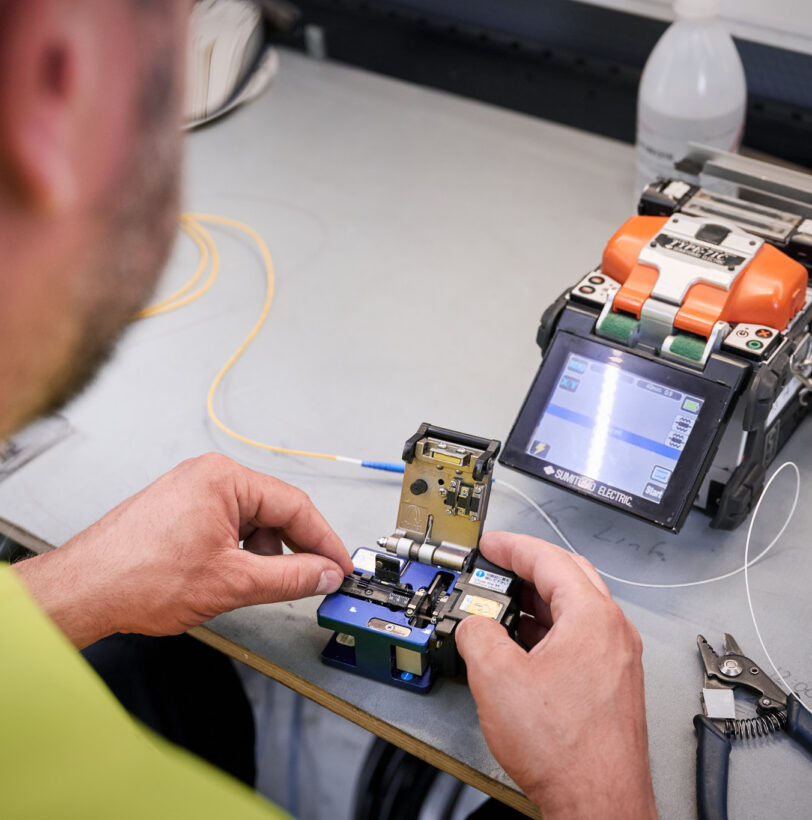 Ein Techniker arbeitet an einer Fusionsspleißmaschine zur Verbindung von Glasfaserkabeln, ein wichtiger Schritt für die Stromnetzinfrastruktur und Mittelspannungsanlagen bei Verteilnetzbetreibern ORES und RESA. Die präzise Handhabung unterstützt den Neubau und Umbau von Trafostationen sowie die Installation schlüsselfertiger Trafostationen und Beton-Fertigstationen in der Energieinfrastruktur Belgien. Werkzeuge und Geräte zur Niederspannungsverteilung und für Transformatoren sind auf dem Arbeitstisch sichtbar.