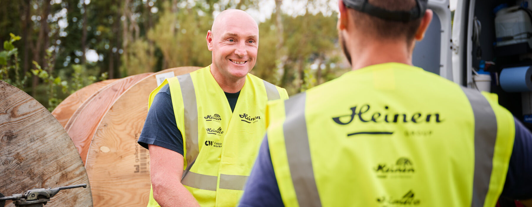 Zwei Handwerker in Warnwesten unterhalten sich vor Transporter und Kabeltrommeln im Außeneinsatz