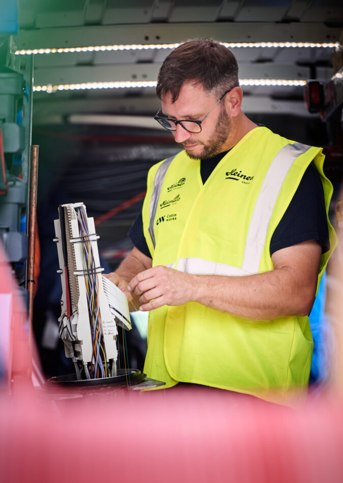 Ein Techniker in Warnweste arbeitet konzentriert an einer Mittelspannungsanlage mit zahlreichen Kabeln und Verbindungen, die Teil der Stromnetzinfrastruktur für Verteilnetzbetreiber ORES und RESA sind. Die Szene symbolisiert den Neubau und Umbau von Trafostationen sowie die Installation von schlüsselfertigen Trafostationen und Beton-Fertigstationen zur Niederspannungsverteilung in der Energieinfrastruktur Belgien. Im Fokus stehen Transformatoren und komplexe Verkabelungen, die für eine zuverlässige Energieversorgung sorgen.
