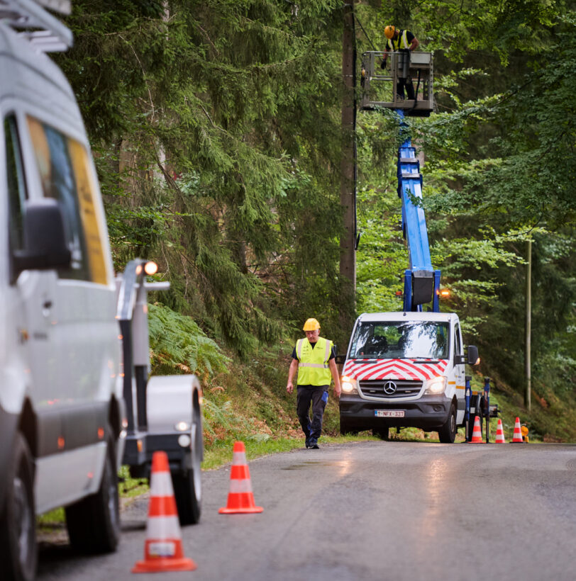 Arbeiter führen Glasfaserarbeiten unter Verkehr in der Provinz Lüttich durch. Ein Techniker steht auf einer Hebebühne an einem Telekommunikationsmast, während ein weiterer Mitarbeiter auf der Straße mit Warnweste und Helm die Baustelle sichert. Zwei Einsatzfahrzeuge sind mit Warnmarkierungen und Verkehrskegeln auf der Straße positioniert. Die Arbeiten umfassen das Spleißen der Glasfaserverbindung sowie das Einziehen und Einblasen von Glasfaserkabeln im Rahmen des Glasfaserausbaus VOO im Hybrid Fibre Coaxial Netz Belgien. Die bidirektionale optische Messung und die Erstellung von einsatzbereiten Glasfaserstrecken mit Dokumentation und Qualitätsmessung sind Teil des Prozesses.