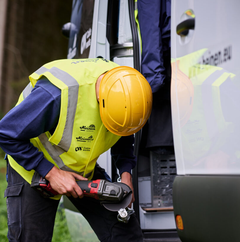 Ein Techniker mit gelbem Schutzhelm und Warnweste führt Glasfaserarbeiten unter Verkehr durch. Er nutzt ein Elektrowerkzeug zum Spleißen der Glasfaserverbindung neben einem weißen Servicefahrzeug. Die Szene zeigt den Glasfaserausbau VOO im Glasfasernetz der Provinz Lüttich in Belgien. Die Arbeiten umfassen das Einziehen und Einblasen von Glasfaserkabeln sowie die bidirektionale optische Messung zur Sicherstellung einsatzbereiter Glasfaserstrecken mit Dokumentation und Qualitätsmessung im Hybrid Fibre Coaxial Netz Belgien.