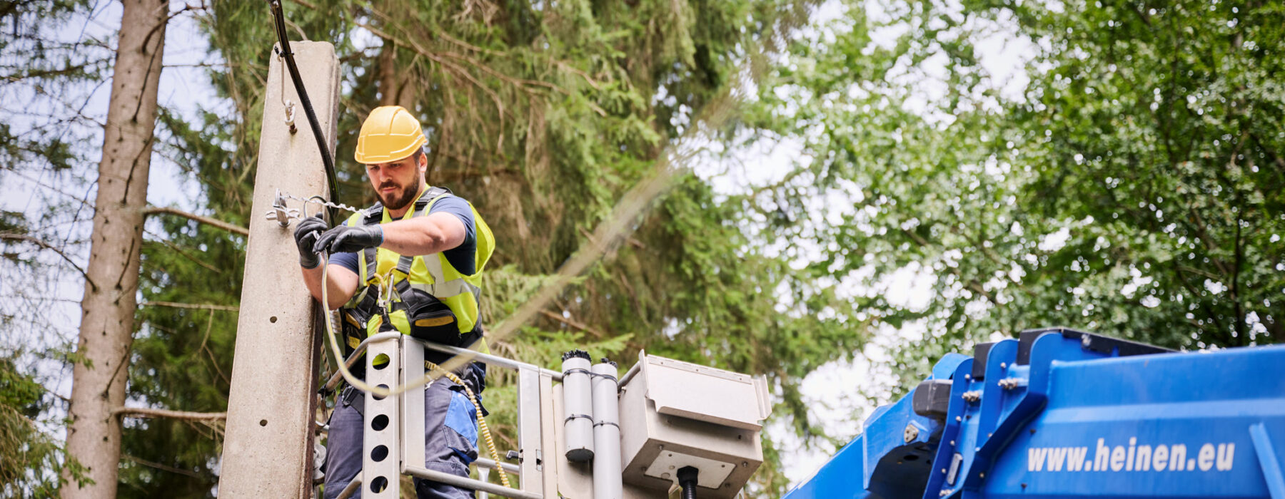 Ein Techniker führt Glasfaserarbeiten unter Verkehr durch, indem er an einem Mast in der Provinz Lüttich eine Glasfaserverbindung spleißt. Er steht in einer Hebebühne und trägt Schutzhelm sowie Sicherheitsweste. Im Rahmen des Glasfaserausbaus VOO werden hier Glasfaserkabel eingezogen und eingeblasen. Die Arbeiten gehören zum Hybrid Fibre Coaxial Belgien Projekt. Die Einsatzbereiten Glasfaserstrecken werden anschließend mit bidirektionaler optischer Messung dokumentiert und auf Qualität geprüft.