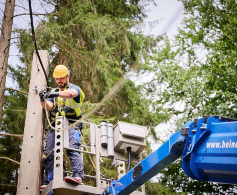 Ein Techniker führt Glasfaserarbeiten unter Verkehr durch, indem er an einem Mast in der Provinz Lüttich eine Glasfaserverbindung spleißt. Er steht in einer Hebebühne und trägt Schutzhelm sowie Sicherheitsweste. Im Rahmen des Glasfaserausbaus VOO werden hier Glasfaserkabel eingezogen und eingeblasen. Die Arbeiten gehören zum Hybrid Fibre Coaxial Belgien Projekt. Die Einsatzbereiten Glasfaserstrecken werden anschließend mit bidirektionaler optischer Messung dokumentiert und auf Qualität geprüft.