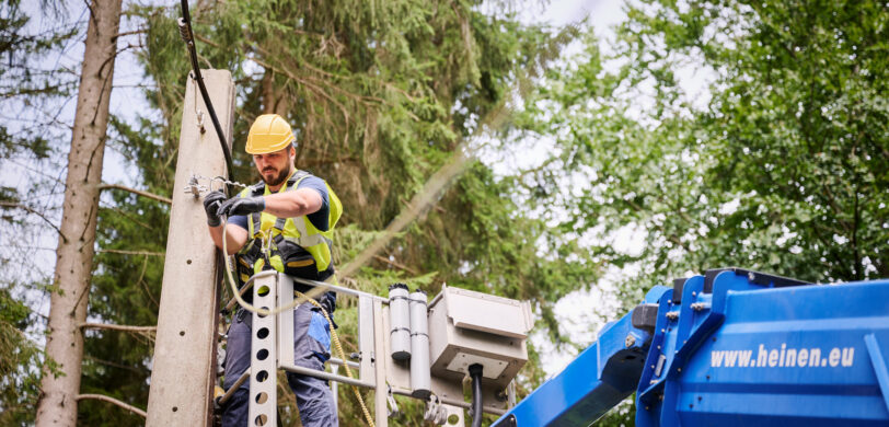 Ein Techniker führt Glasfaserarbeiten unter Verkehr durch, indem er an einem Mast in der Provinz Lüttich eine Glasfaserverbindung spleißt. Er steht in einer Hebebühne und trägt Schutzhelm sowie Sicherheitsweste. Im Rahmen des Glasfaserausbaus VOO werden hier Glasfaserkabel eingezogen und eingeblasen. Die Arbeiten gehören zum Hybrid Fibre Coaxial Belgien Projekt. Die Einsatzbereiten Glasfaserstrecken werden anschließend mit bidirektionaler optischer Messung dokumentiert und auf Qualität geprüft.