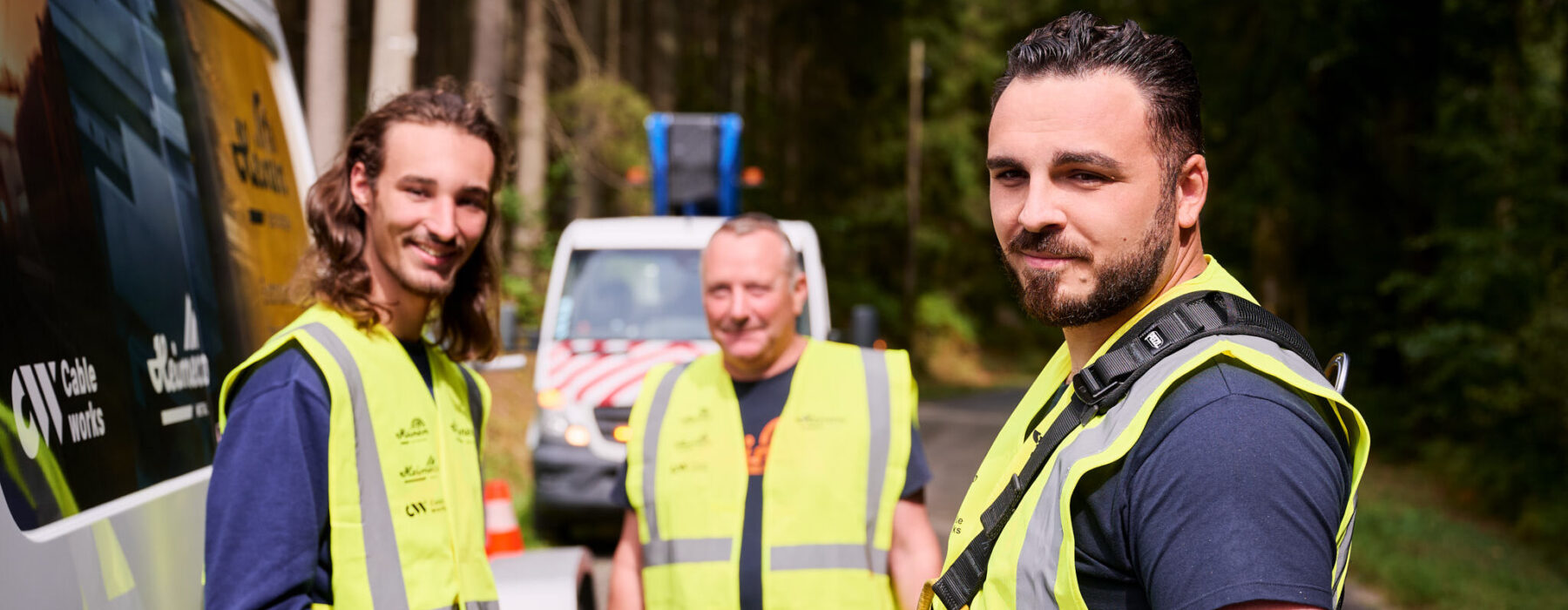 Drei Bauarbeiter in reflektierenden Warnwesten stehen vor einem Einsatzfahrzeug auf einer Waldstraße. Sie wirken als zentraler Ansprechpartner und vereinen Kompetenz durch ihre vielseitige und lösungsorientierte Beratung. Die Szene ist durchdacht und transparent gestaltet, um professionelle Zusammenarbeit und Sicherheit zu vermitteln.