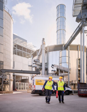 Zwei Elektriker mit Schutzhelmen und Warnwesten gehen vor einem industriellen Sägewerk in Vielsalm, ausgestattet mit einem Blockheizkraftwerk für Industrie und moderner elektrotechnischer Ausstattung für Holzindustrie. Im Hintergrund sind große Silos, Rohrleitungen und eine Kopfstation sichtbar, die Teil der industriellen Energieverteilung sind. Ein Firmenfahrzeug steht bereit, während die Integration des BHKW in das bestehende Mittelspannungsnetz und die Einspeisung in das öffentliche Mittelspannungsnetz durch Glasfaser Leitungen Industrie unterstützt wird. Die Anlage entspricht einem detaillierten Lastenheft und umfasst auch eine industrielle Photovoltaikanlage zur nachhaltigen Energieversorgung. Die Elektrotechnik in Industriehallen wird hier praxisnah umgesetzt.