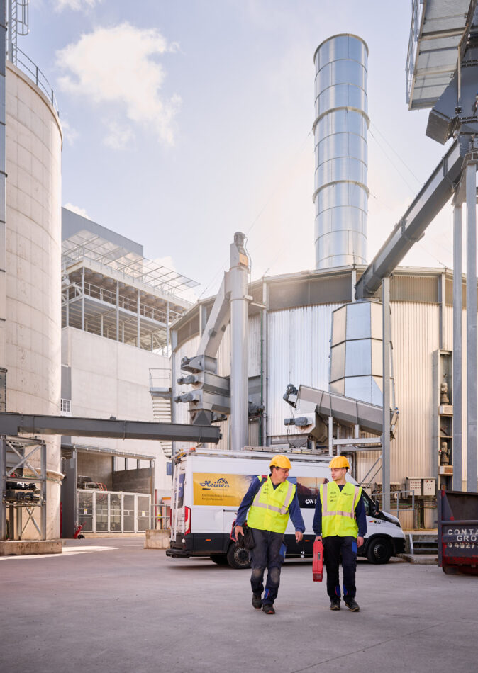 Zwei Elektriker mit Schutzhelmen und Warnwesten gehen vor einem industriellen Sägewerk in Vielsalm, ausgestattet mit einem Blockheizkraftwerk für Industrie und moderner elektrotechnischer Ausstattung für Holzindustrie. Im Hintergrund sind große Silos, Rohrleitungen und eine Kopfstation sichtbar, die Teil der industriellen Energieverteilung sind. Ein Firmenfahrzeug steht bereit, während die Integration des BHKW in das bestehende Mittelspannungsnetz und die Einspeisung in das öffentliche Mittelspannungsnetz durch Glasfaser Leitungen Industrie unterstützt wird. Die Anlage entspricht einem detaillierten Lastenheft und umfasst auch eine industrielle Photovoltaikanlage zur nachhaltigen Energieversorgung. Die Elektrotechnik in Industriehallen wird hier praxisnah umgesetzt.