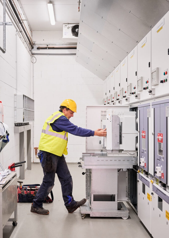 Ein Techniker in Schutzhelm und Warnweste arbeitet an einer elektrotechnischen Schaltanlage in einer Industriehalle. Die Anlage umfasst Schaltschränke und Mittelspannungskomponenten, die für die Integration eines Blockheizkraftwerks (BHKW) in das bestehende Mittelspannungsnetz konzipiert sind. Im Raum sind außerdem Löschgasflaschen sichtbar, die zur Sicherheit der elektrotechnischen Ausstattung für die Holzindustrie beitragen. Die Installation entspricht einem detaillierten Lastenheft und ermöglicht die Einspeisung in das öffentliche Mittelspannungsnetz. Die moderne Infrastruktur unterstützt zudem Glasfaser Leitungen für Industrie und die Anbindung einer industriellen Photovoltaikanlage. Die Kopfstationen und die industrielle Energieverteilung sind Teil der Elektrotechnik im Sägewerk IBV Vielsalm.
