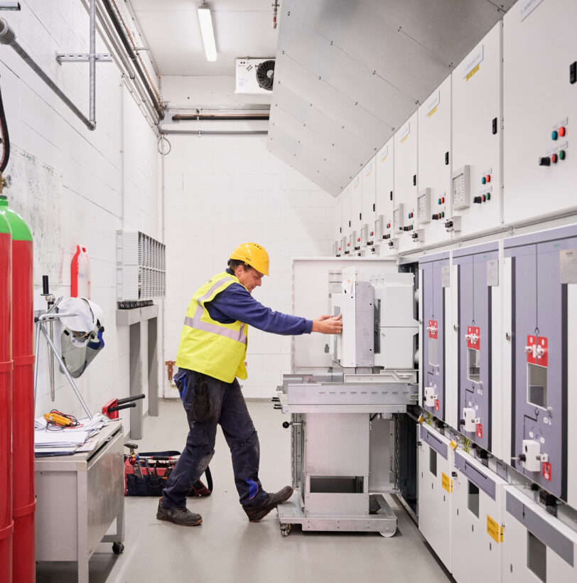 Ein Techniker in Schutzhelm und Warnweste arbeitet an einer elektrotechnischen Schaltanlage in einer Industriehalle. Die Anlage umfasst Schaltschränke und Mittelspannungskomponenten, die für die Integration eines Blockheizkraftwerks (BHKW) in das bestehende Mittelspannungsnetz konzipiert sind. Im Raum sind außerdem Löschgasflaschen sichtbar, die zur Sicherheit der elektrotechnischen Ausstattung für die Holzindustrie beitragen. Die Installation entspricht einem detaillierten Lastenheft und ermöglicht die Einspeisung in das öffentliche Mittelspannungsnetz. Die moderne Infrastruktur unterstützt zudem Glasfaser Leitungen für Industrie und die Anbindung einer industriellen Photovoltaikanlage. Die Kopfstationen und die industrielle Energieverteilung sind Teil der Elektrotechnik im Sägewerk IBV Vielsalm.