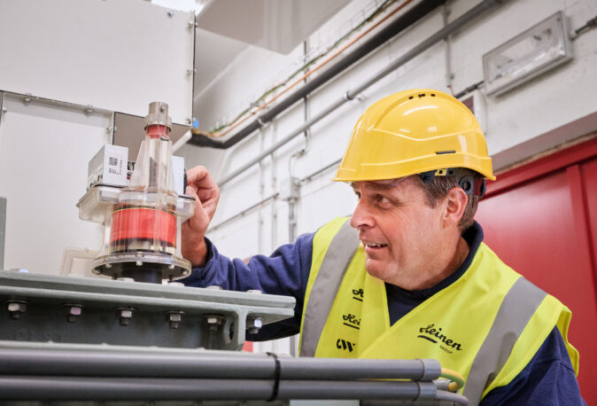 Ein Techniker mit gelbem Schutzhelm und Warnweste inspiziert eine elektrotechnische Anlage in einer Industriehalle. Die Installation zeigt Komponenten zur Integration eines Blockheizkraftwerks (BHKW) in das bestehende Mittelspannungsnetz, inklusive Kopfstationen und industrieller Energieverteilung. Die Elektrotechnik im Sägewerk IBV Vielsalm umfasst auch Glasfaser Leitungen für Industrie und eine industrielle Photovoltaikanlage, die gemäß Lastenheft für die Einspeisung in das öffentliche Mittelspannungsnetz ausgelegt ist. Die elektrotechnische Ausstattung für die Holzindustrie wird hier praxisnah umgesetzt.