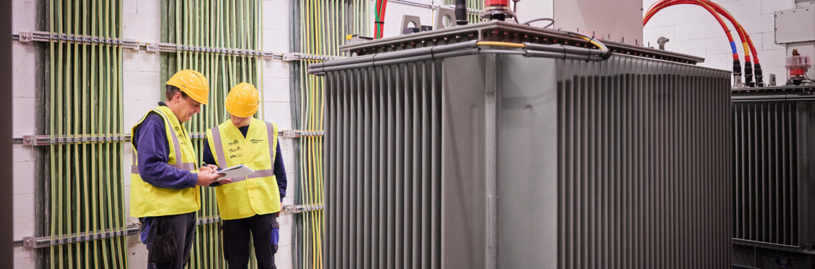 Zwei Techniker mit gelben Schutzhelmen und Warnwesten überprüfen ein Lastenheft in einem Raum mit umfangreicher elektrotechnischer Ausstattung für Holzindustrie. Im Vordergrund steht ein großer Transformator mit dicken roten und grünen Kabeln, die in Kopfstationen verlaufen. Die Installation zeigt die Integration eines Blockheizkraftwerks (BHKW) in das bestehende Mittelspannungsnetz zur Einspeisung in das öffentliche Mittelspannungsnetz. Im Hintergrund sind geordnete Glasfaser Leitungen Industrie an der Wand befestigt, die eine industrielle Energieverteilung und Elektrotechnik in Industriehallen gewährleisten. Die Szene repräsentiert Elektrotechnik im Sägewerk IBV Vielsalm mit Fokus auf industrielle Photovoltaikanlage und moderne Energieversorgung.