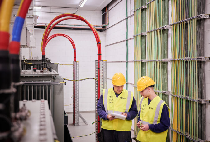 Zwei Elektriker in Schutzhelmen und Warnwesten prüfen und dokumentieren die elektrotechnische Ausstattung in einer Industriehalle eines Sägewerks. Im Hintergrund sind umfangreiche Glasfaser Leitungen Industrie und Kabelstränge zu sehen, die in Kopfstationen organisiert sind. Die Installation zeigt die Integration eines Blockheizkraftwerks für Industrie in das bestehende Mittelspannungsnetz mit Einspeisung in das öffentliche Mittelspannungsnetz. Die industrielle Energieverteilung und die Umsetzung eines Lastenhefts für Elektrotechnik in Industriehallen sind hier exemplarisch dargestellt. Die Elektrotechnik Sägewerk IBV Vielsalm steht für moderne Lösungen in der Holzindustrie, inklusive industrieller Photovoltaikanlage und effizienter Energieverteilung.