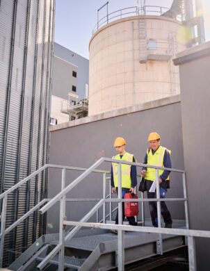 Zwei Techniker mit Schutzhelmen und Warnwesten gehen auf einer Metalltreppe in einem Industriekomplex, der zur Elektrotechnik im Sägewerk IBV Vielsalm gehört. Im Hintergrund sind große industrielle Anlagen und Silos zu sehen, die Teil der elektrotechnischen Ausstattung für die Holzindustrie sind. Die Szene symbolisiert die Integration eines Blockheizkraftwerks (BHKW) in das bestehende Mittelspannungsnetz sowie die industrielle Energieverteilung. Die Installation umfasst Glasfaser Leitungen für die Industrie, Kopfstationen und eine industrielle Photovoltaikanlage zur Einspeisung in das öffentliche Mittelspannungsnetz gemäß dem Lastenheft.