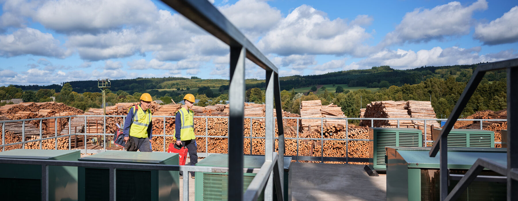 Zwei Mitarbeiter mit Schutzhelmen und Warnwesten gehen auf einer Plattform zwischen elektrotechnischen Anlagen in einem Sägewerk in Vielsalm. Im Hintergrund sind große Holzstapel zu sehen, die auf die Holzindustrie hinweisen. Die Installation zeigt die elektrotechnische Ausstattung für Holzindustrie, inklusive Kopfstationen und Integration eines Blockheizkraftwerks (BHKW) in das bestehende Mittelspannungsnetz. Die Szene symbolisiert die industrielle Energieverteilung und die Einspeisung in das öffentliche Mittelspannungsnetz. Die Umgebung deutet auf Elektrotechnik in Industriehallen und die Nutzung von Glasfaser Leitungen in der Industrie hin. Das Lastenheft für die Umsetzung einer industriellen Photovoltaikanlage ist Teil des nachhaltigen Energiekonzepts.