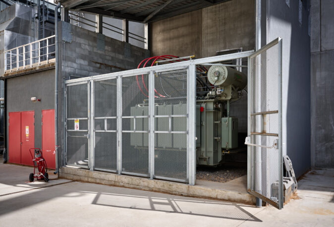 Das Bild zeigt die elektrotechnische Ausstattung eines Blockheizkraftwerks (BHKW) in einem Industriegebäude eines Sägewerks in Vielsalm. Im Vordergrund ist ein großer Transformator hinter einem Metallgitterzaun mit offener Tür zu sehen, der zur Integration des BHKW in das bestehende Mittelspannungsnetz dient. Im Hintergrund sind Industriehallen mit Betonwänden und Lüftungssystemen sichtbar. Die Installation entspricht den Anforderungen eines Lastenhefts für die Einspeisung in das öffentliche Mittelspannungsnetz. Die Anlage ist Teil der industriellen Energieverteilung und umfasst auch Glasfaser Leitungen für die Industriekommunikation sowie Kopfstationen. Zusätzlich ist die industrielle Photovoltaikanlage für nachhaltige Energieerzeugung in der Holzindustrie berücksichtigt. Die Elektrotechnik im Sägewerk IBV Vielsalm gewährleistet eine zuverlässige Versorgung und Steuerung der Energieflüsse in der Industriehalle.