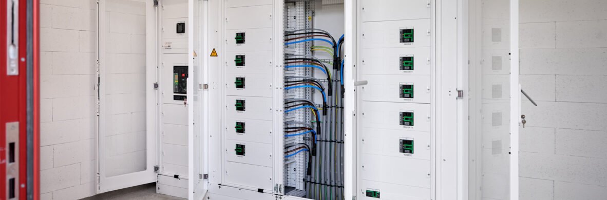 Weiße elektrotechnische Schaltschränke mit geöffneten Türen in einem Raum mit Betonboden und weißen Wänden, ausgestattet für die industrielle Energieverteilung. Die Installation zeigt Kabel und Sicherungen, die für die Integration eines Blockheizkraftwerks (BHKW) in das bestehende Mittelspannungsnetz vorbereitet sind. Diese elektrotechnische Ausstattung für Holzindustrie und Industriehallen entspricht dem Lastenheft für Elektrotechnik im Sägewerk IBV Vielsalm. Die Schaltschränke sind Teil der Kopfstationen und ermöglichen die Einspeisung in das öffentliche Mittelspannungsnetz. Die Anlage unterstützt auch Glasfaser Leitungen für Industrie und ist kompatibel mit industriellen Photovoltaikanlagen.