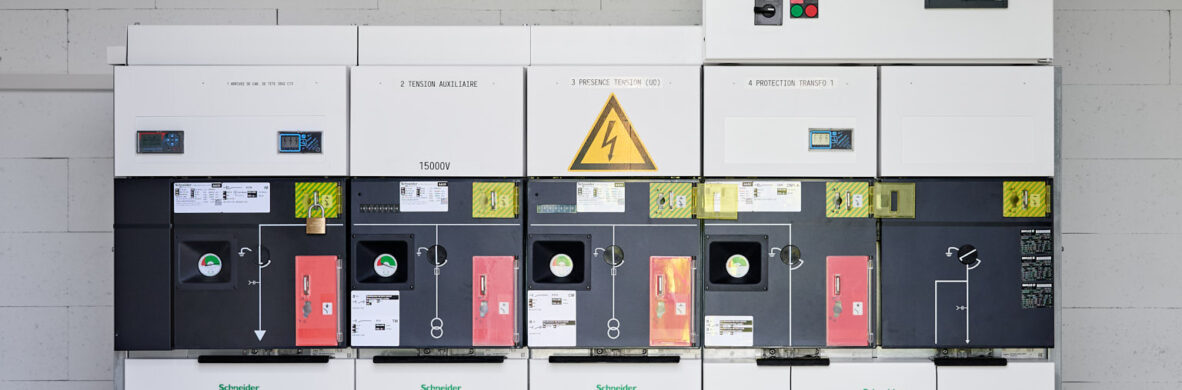Schaltschrank der Marke Schneider Electric in einer Industriehalle, Teil der elektrotechnischen Ausstattung für Holzindustrie im Sägewerk IBV Vielsalm. Das Gerät dient der industriellen Energieverteilung und ist für die Integration eines Blockheizkraftwerks (BHKW) in das bestehende Mittelspannungsnetz ausgelegt. Die Anlage entspricht dem Lastenheft für die Einspeisung in das öffentliche Mittelspannungsnetz und unterstützt Kopfstationen sowie Glasfaser Leitungen in der Industrie. Die Installation ist Teil einer industriellen Photovoltaikanlage und gewährleistet Elektrotechnik in Industriehallen.