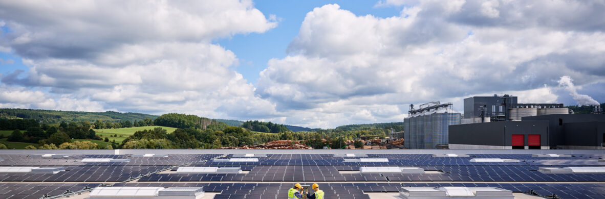 Techniker prüfen industrielle Photovoltaikanlage auf Sägewerksdach mit Industriehallen im Hintergrund