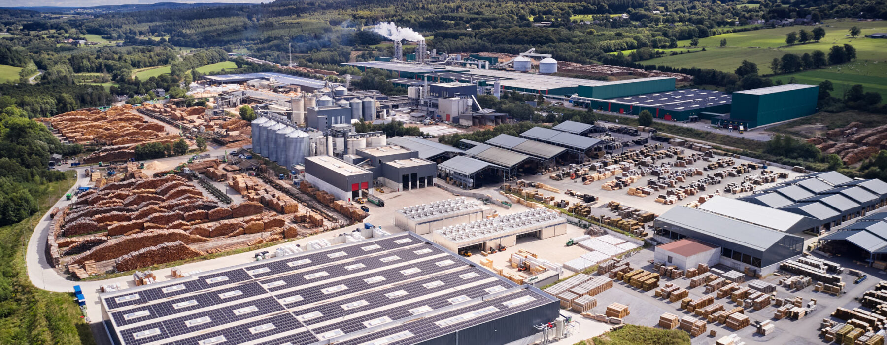 Luftaufnahme eines großen Sägewerks mit umfangreicher Elektrotechnik in Vielsalm. Im Vordergrund ist eine industrielle Photovoltaikanlage auf dem Dach einer Industriehalle zu sehen, die zur nachhaltigen Energieversorgung beiträgt. Das Werk ist mit Glasfaser Leitungen für Industriekommunikation ausgestattet. Im Hintergrund sind mehrere Lagerflächen mit Holzstapeln und Silos zu erkennen. Die elektrotechnische Ausstattung für Holzindustrie umfasst Kopfstationen und die Integration eines Blockheizkraftwerks (BHKW) in das bestehende Mittelspannungsnetz. Die Einspeisung in das öffentliche Mittelspannungsnetz erfolgt nach einem detaillierten Lastenheft. Das Bild zeigt exemplarisch die industrielle Energieverteilung und Elektrotechnik in Industriehallen eines modernen Sägewerks der IBV Vielsalm.