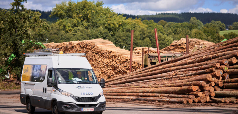 Ein weißer Iveco Transporter mit der Aufschrift Heinen fährt auf dem Gelände eines Sägewerks in Vielsalm vorbei an großen Holzstapeln. Das Bild zeigt die Elektrotechnik im Sägewerk IBV Vielsalm, inklusive der elektrotechnischen Ausstattung für Holzindustrie und der Integration eines Blockheizkraftwerks (BHKW) in das bestehende Mittelspannungsnetz. Im Hintergrund sind Kopfstationen und Glasfaser Leitungen für die Industrie sichtbar. Die industrielle Energieverteilung und die Einspeisung in das öffentliche Mittelspannungsnetz sind Teil des Lastenhefts für die industrielle Photovoltaikanlage und Elektrotechnik in Industriehallen.