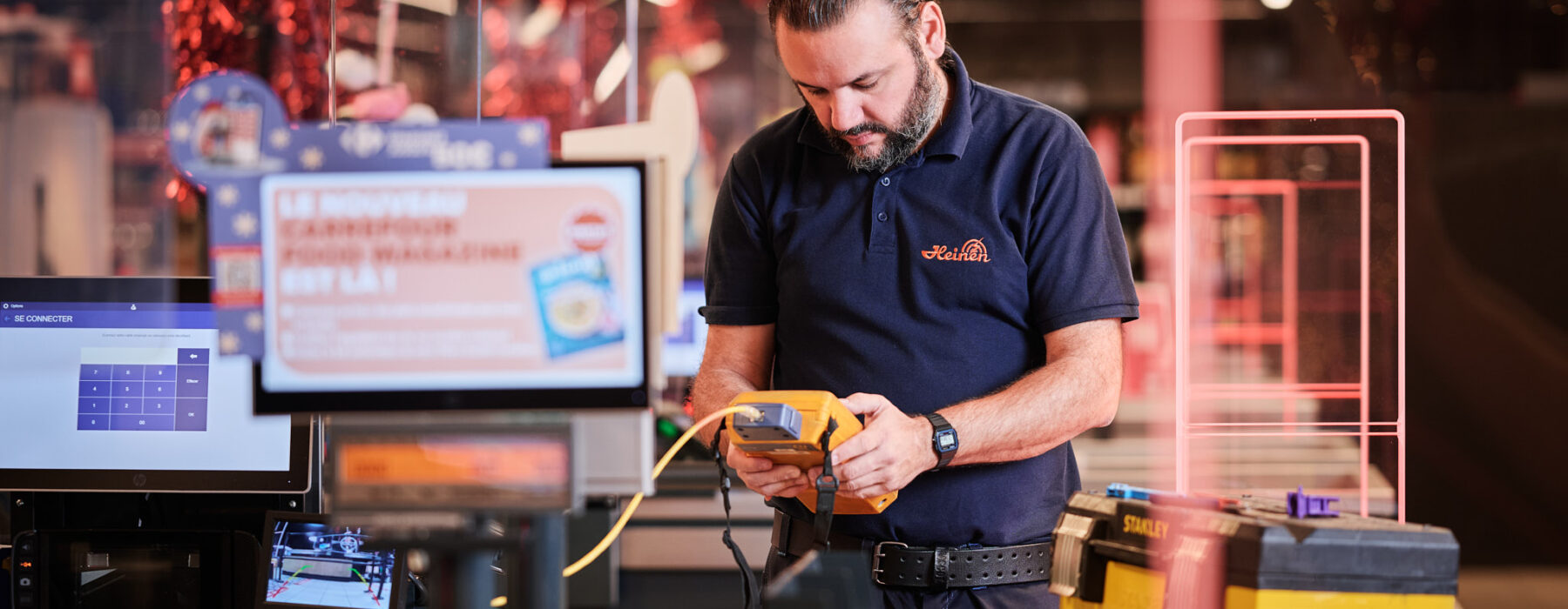 Ein Techniker führt Elektroinstallationen im Carrefour Supermarkt Belgien durch. Er arbeitet an Schaltanlagen und einer Niederspannungsanlage, um die Not- und Sicherheitsbeleuchtung im Einzelhandel zu gewährleisten. Im Vordergrund sind Metallelemente des Geschäfts sowie ein Werkzeugkasten sichtbar. Die Arbeiten tragen zum Einbruchschutz für Supermärkte und zur Einrichtung von Kommunikationsnetzen im Einzelhandel bei.