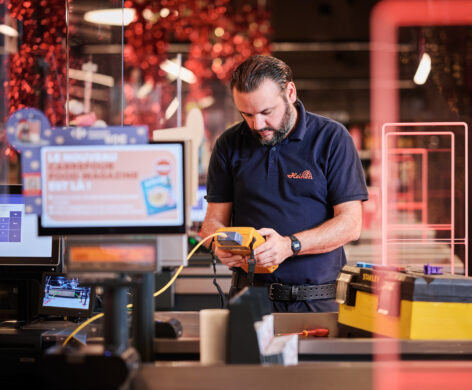 Ein Techniker führt Elektroinstallationen im Carrefour Supermarkt Belgien durch. Er arbeitet an Schaltanlagen und einer Niederspannungsanlage, um die Not- und Sicherheitsbeleuchtung im Einzelhandel zu gewährleisten. Im Vordergrund sind Metallelemente des Geschäfts sowie ein Werkzeugkasten sichtbar. Die Arbeiten tragen zum Einbruchschutz für Supermärkte und zur Einrichtung von Kommunikationsnetzen im Einzelhandel bei.