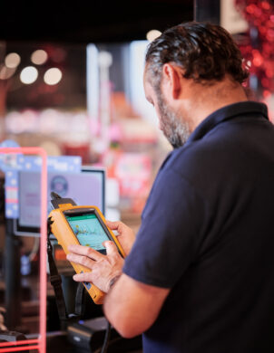 Ein Techniker im Carrefour Supermarkt Belgien überprüft mit einem Messgerät die Kommunikationsnetze im Einzelhandel. Die Szene zeigt moderne Elektroinstallationen im Supermarkt, die Schaltanlagen und Niederspannungsanlage umfassen. Im Hintergrund sind Metallelemente des Geschäfts sichtbar, die zum Einbruchschutz für Supermärkte beitragen. Die Not- und Sicherheitsbeleuchtung im Einzelhandel ist ebenfalls Teil der Installation, die für einen sicheren Betrieb sorgt.